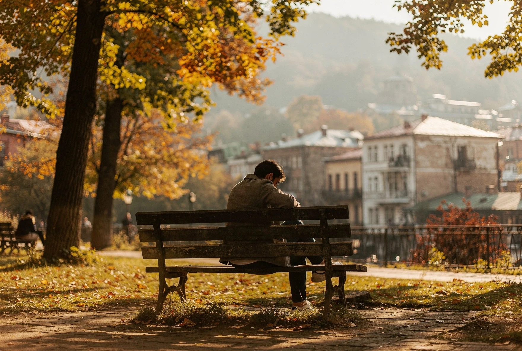 Person sitting alone on a park bench in autumn with Tbilisi buildings in the background