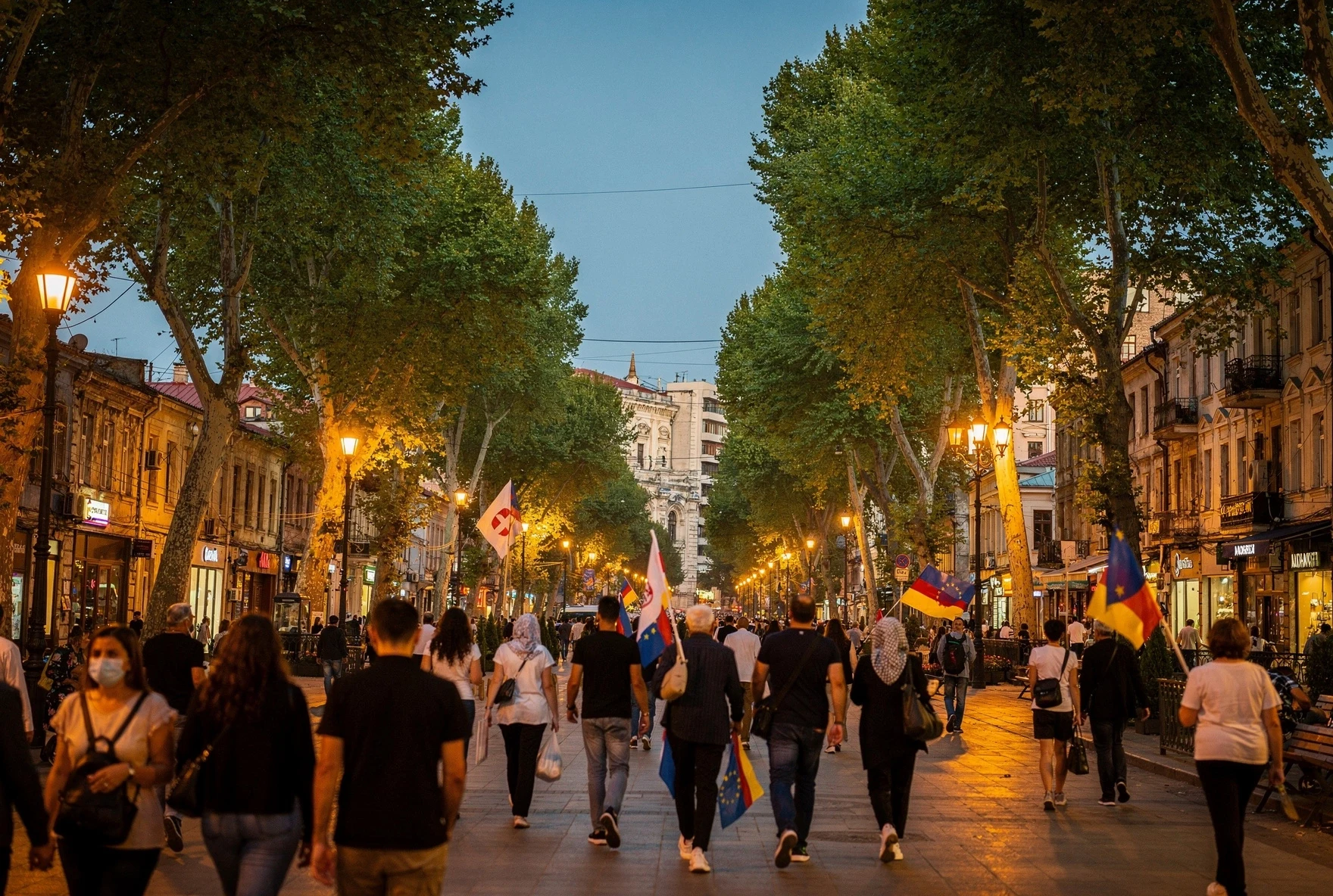 Peaceful crowd walking along a tree-lined boulevard in Tbilisi at dusk with flags