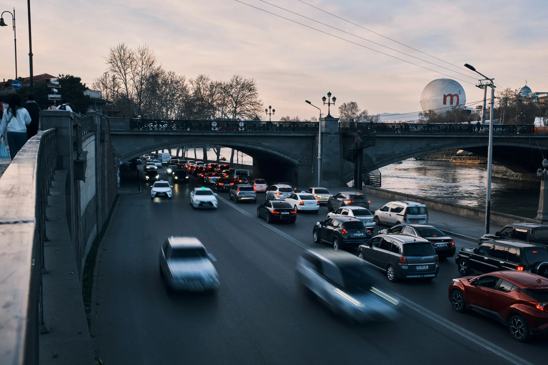 Evening rush hour traffic on a Tbilisi road passing under a stone bridge by the Kura River